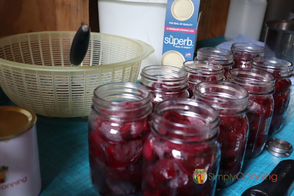 Table with jars of home canned beets in the foreground and a yellow plastic strainer, all sitting around a box of regular size superb canning lids.