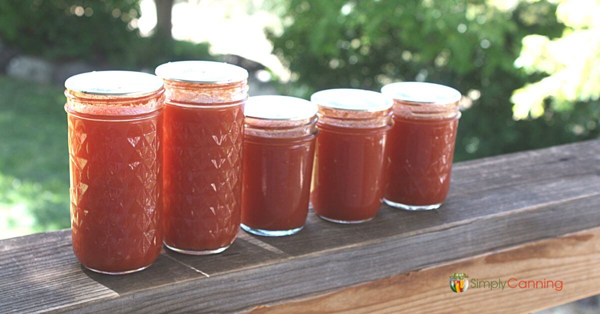 5 small jars of tomato juice lined on on the deck railing. 