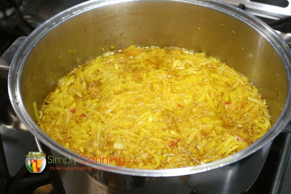 Pot of shredded sweet zucchini relish being cooked in a large pot, yellow color with bits of red onions.